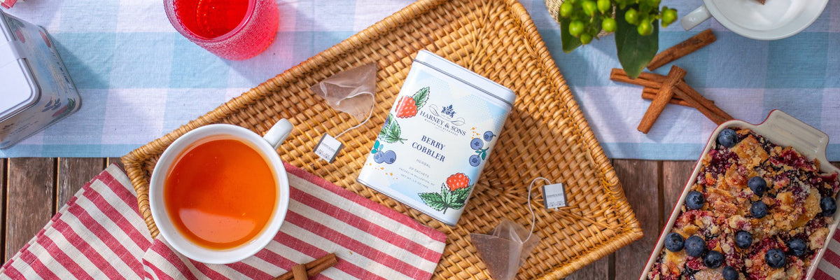 Tea cup with a tea bag, Berry Cobbler tea tin, and snacks on a tablecloth.
