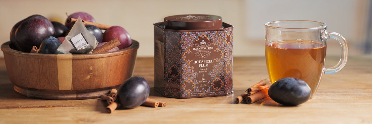 Harney & Sons Hot Spiced Plum tin with a cup of tea, a bowl of plums, and cinnamon sticks