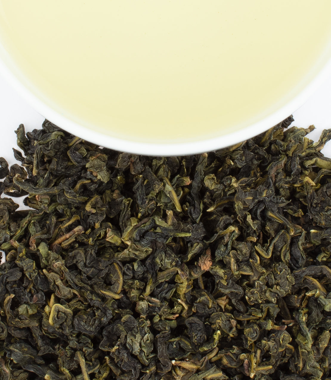 Dried Qing Ti Quan Yin tea leaves with a cup of pale yellow brewed tea above them.