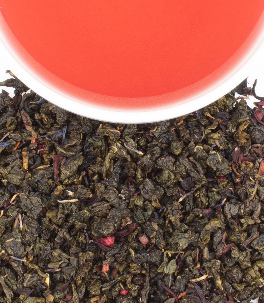 Green curled tea leaves and red and blue flowers with a cup of red tea in the background