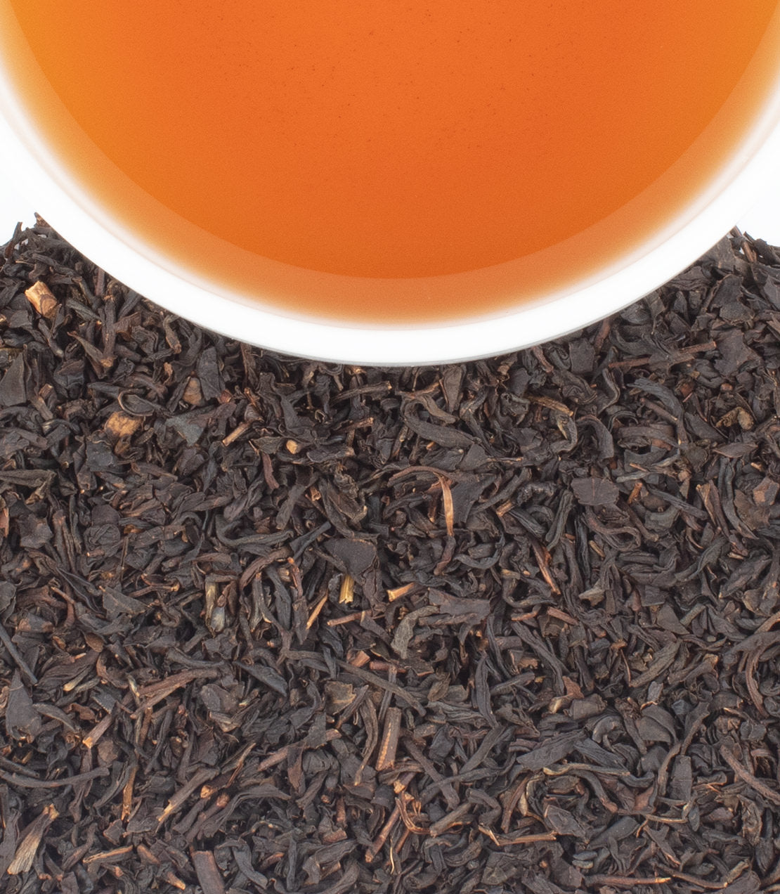 Close-up of loose dark tea leaves with a white cup rim and amber tea visible at the top, photographed against a clean white background.