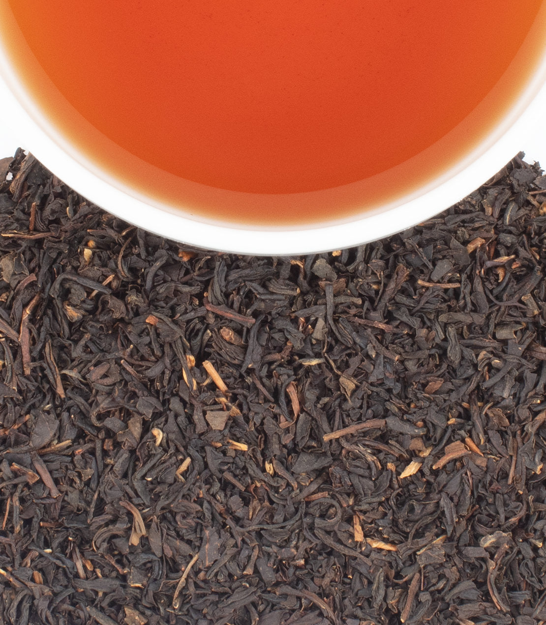 Close-up of loose dark tea leaves with a white cup rim and amber tea liquid at the top, photographed against a clean white background.