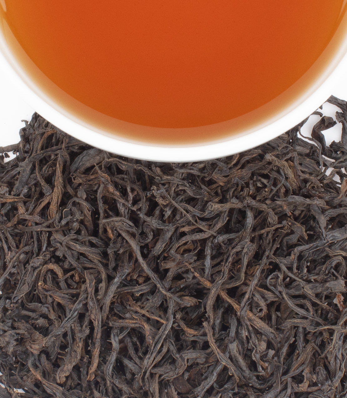 A white cup filled with amber tea sits atop a pile of dark, twisted tea leaves.