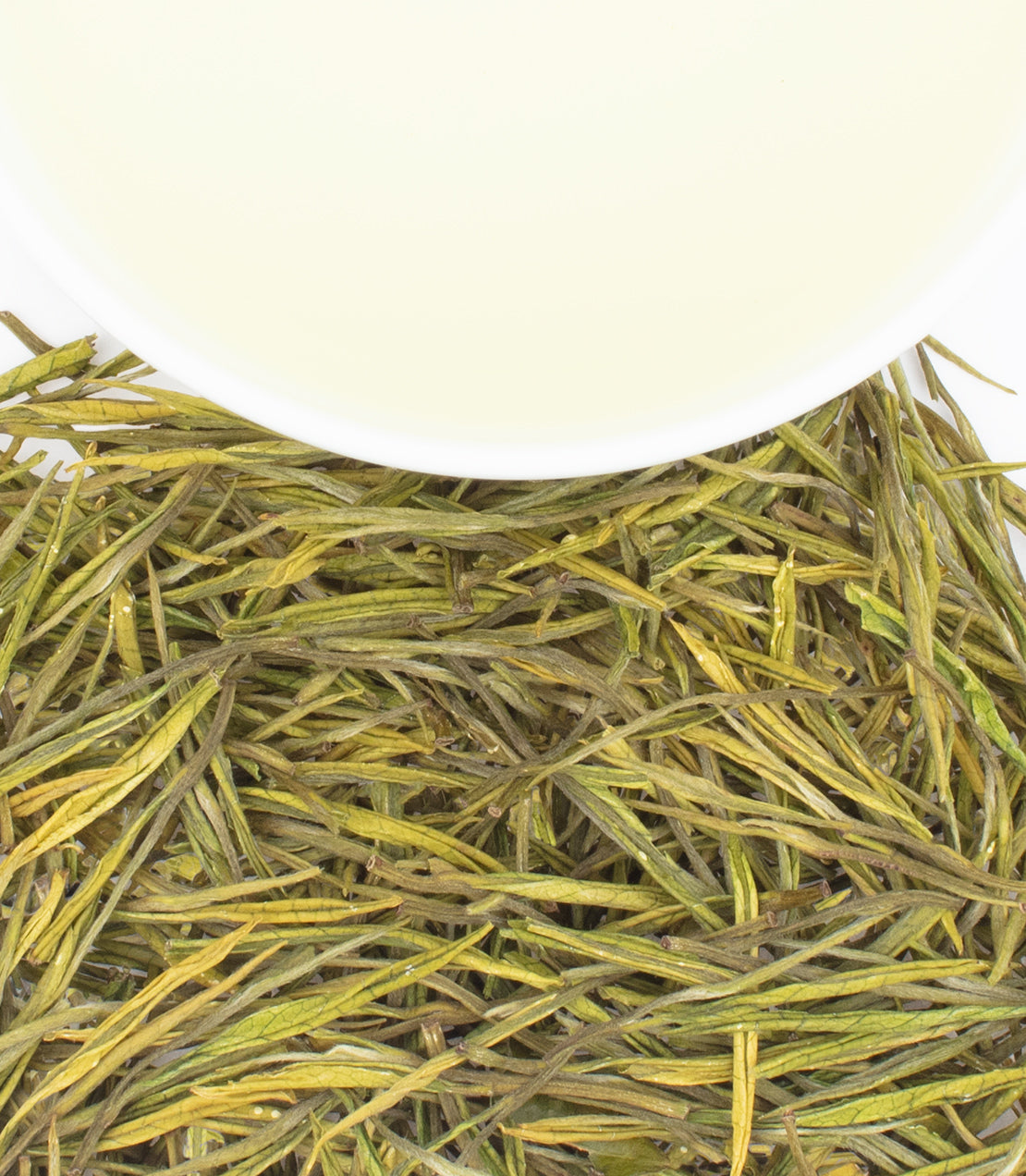 A cup of pale tea rests above a pile of slender, green tea leaves.