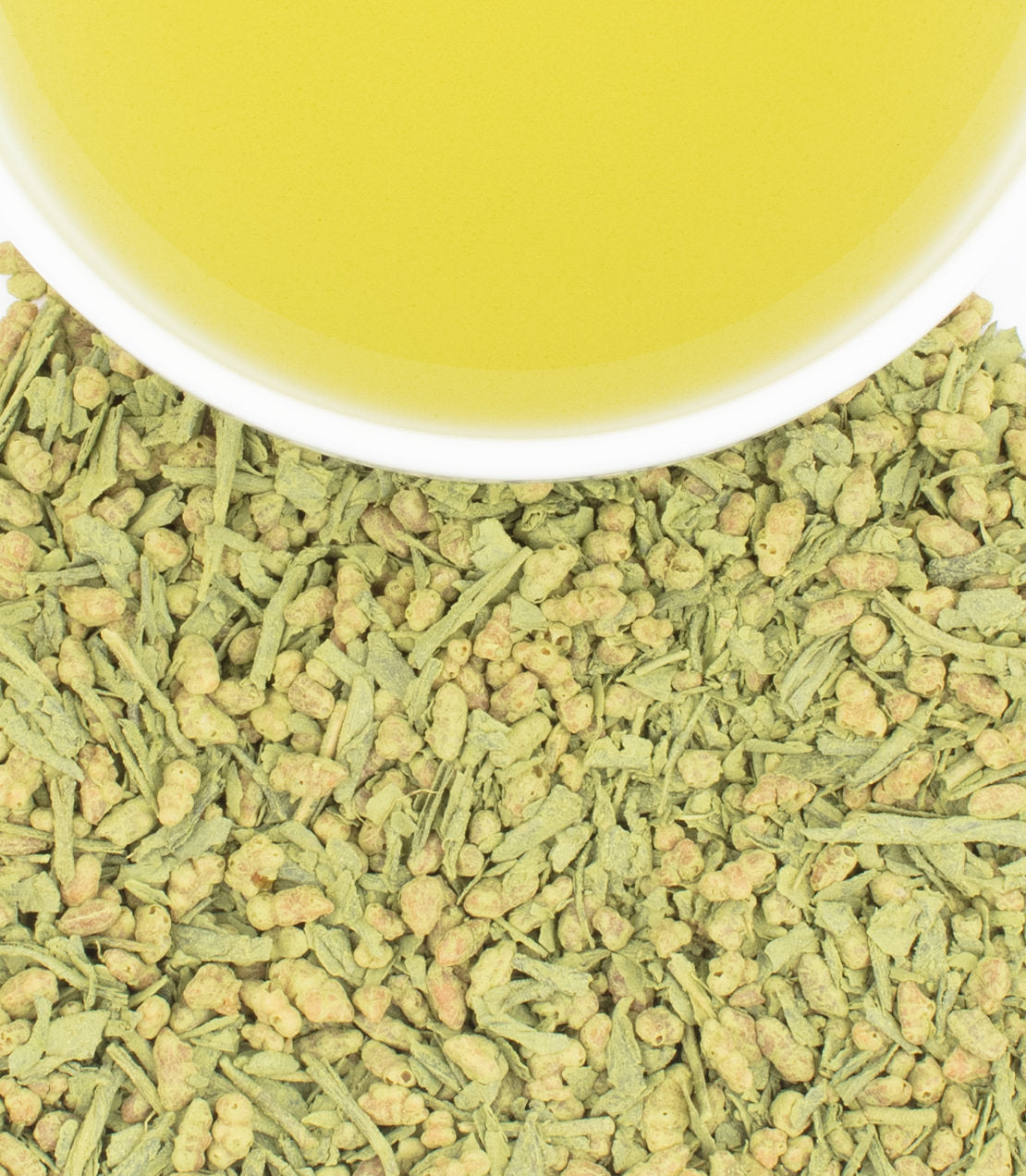 A cup of pale yellow tea rests atop a bed of loose green tea leaves and rice coated in matcha