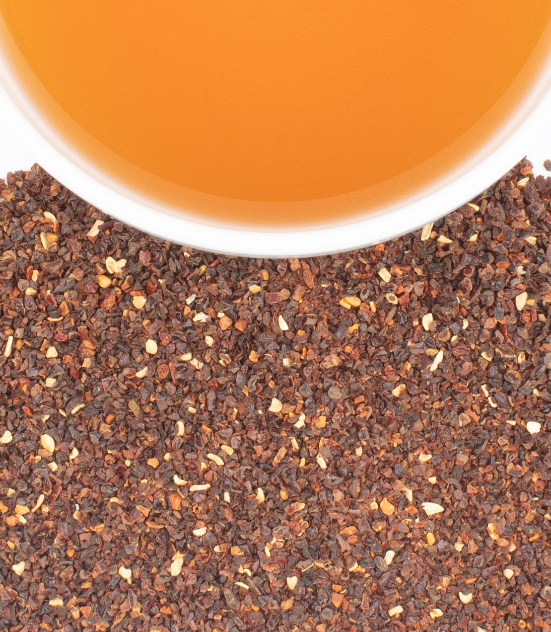 A white cup filled with amber tea sits above a pile of darker, crushed rose hips and small light-colored pieces.