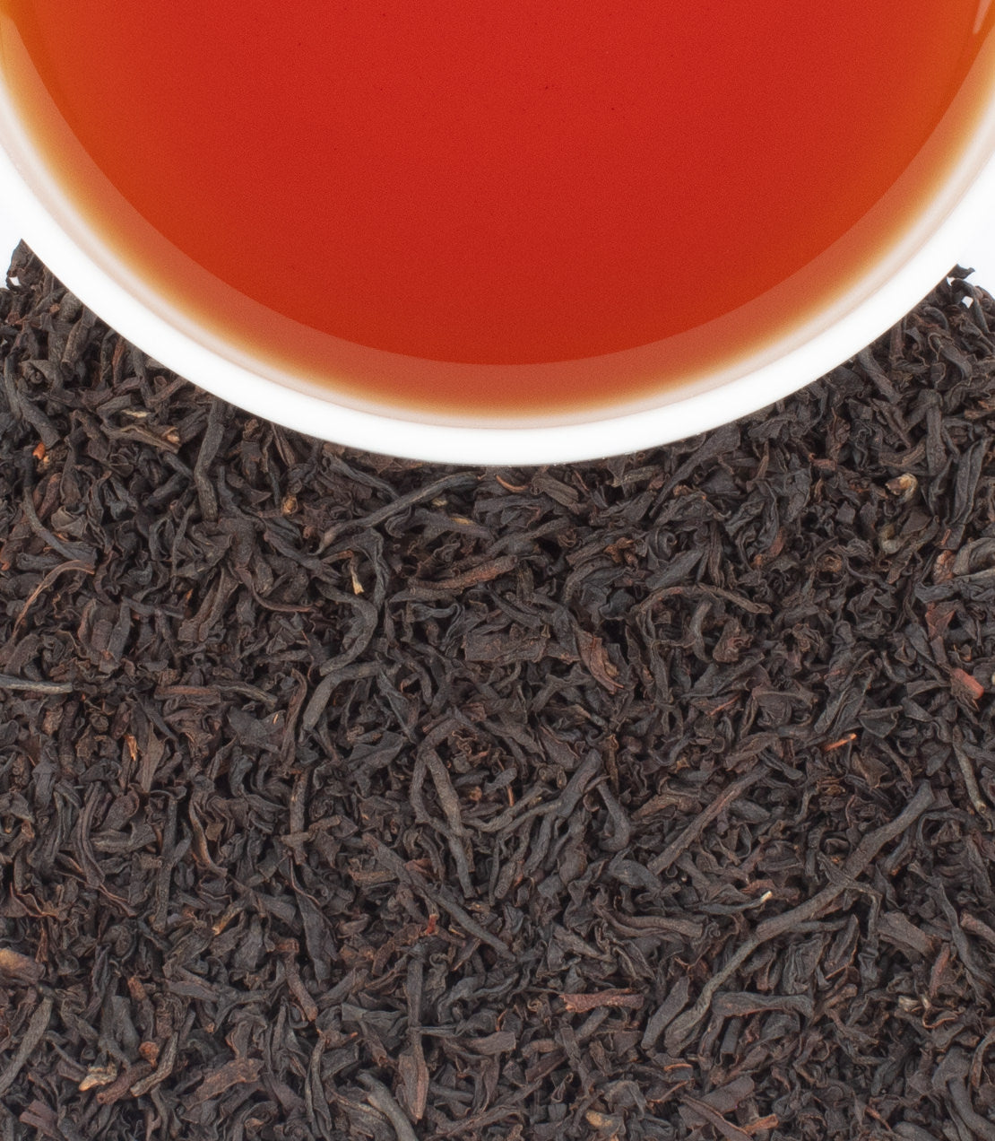 A white cup filled with amber tea sits above a pile of dark, curly black tea leaves.