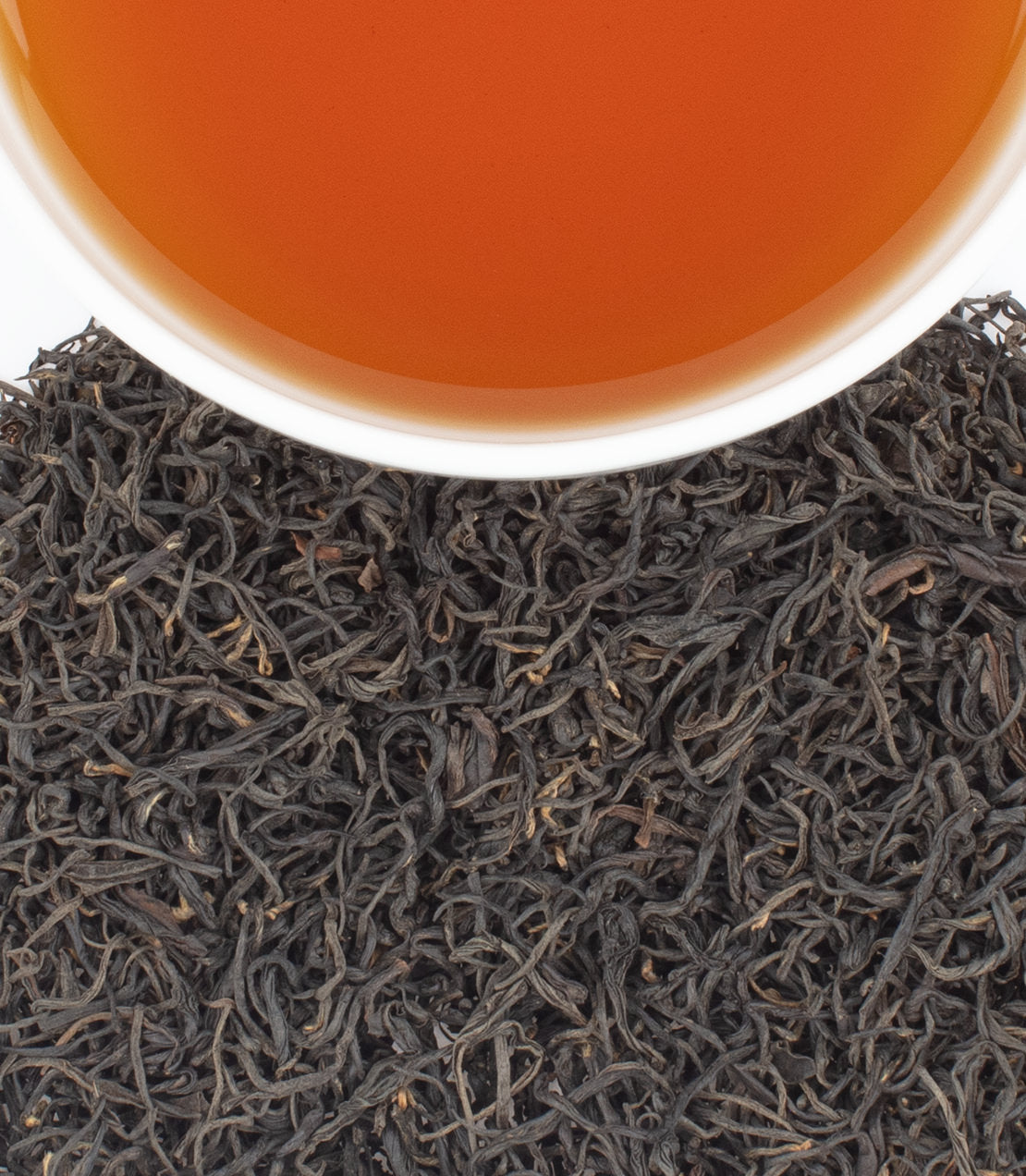 A cup of amber tea sits above a pile of dark, curly loose-leaf tea.