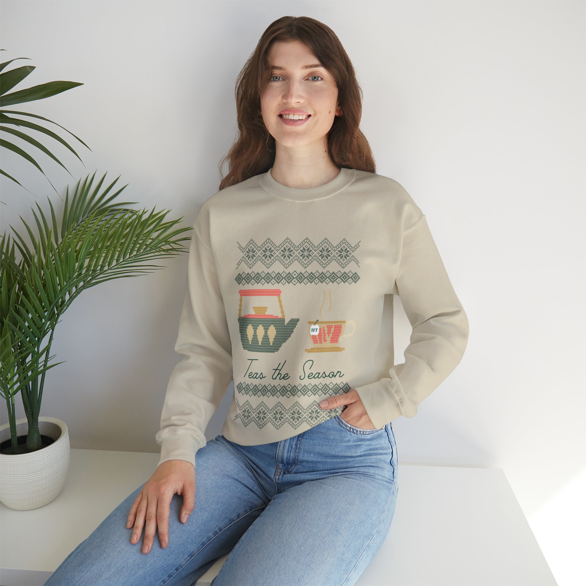 Woman wearing a beige holiday sweater with "Teas The Season" and festive tea-themed graphics, seated by a plant.