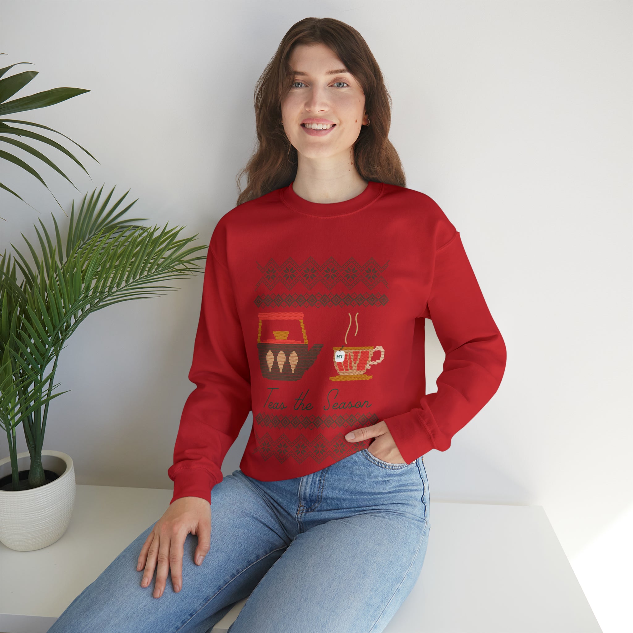 Model wearing a red holiday sweater featuring a festive tea design, sitting against a light background with a plant.