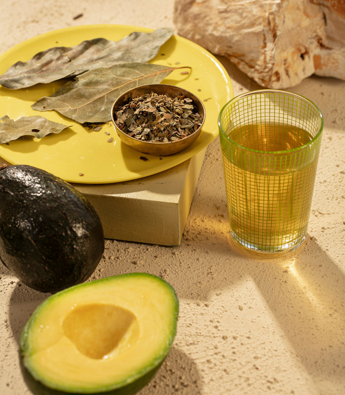 Fresh avocado and avocado leaves on a yellow plate, with a glass of herbal tea and a bowl of dried herbs.