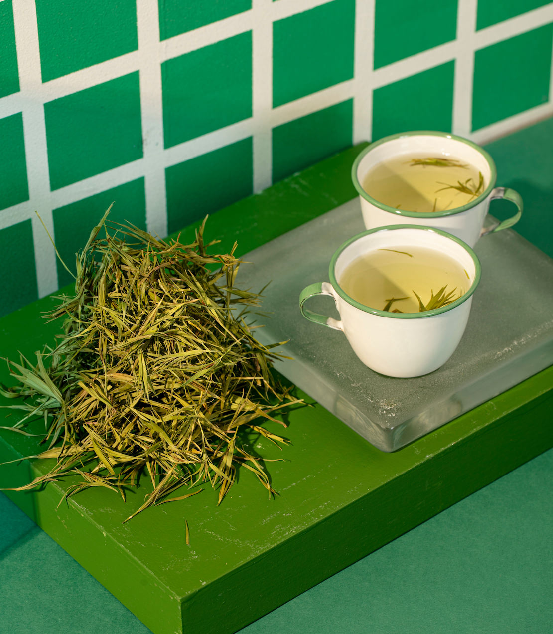 Two cups of bamboo tea sit on a green surface next to a pile of fresh bamboo leaves, against a patterned background.