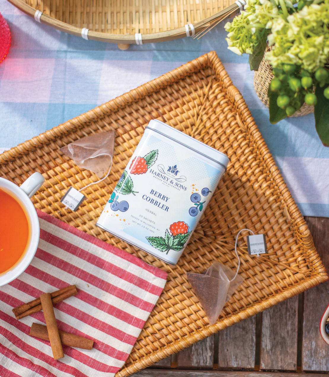 Tin of 20 Berry Cobbler tea sachets on a woven tray, surrounded by a cup of tea and decorative elements.