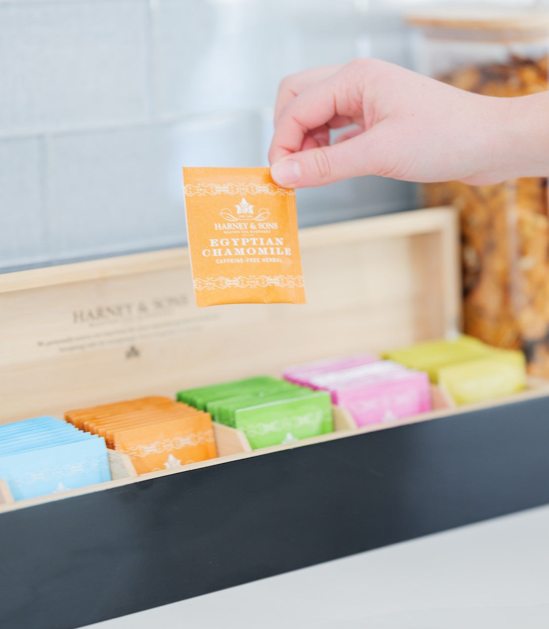 Hand holding a Harney & Sons tea bag over a wooden box of assorted tea bags.