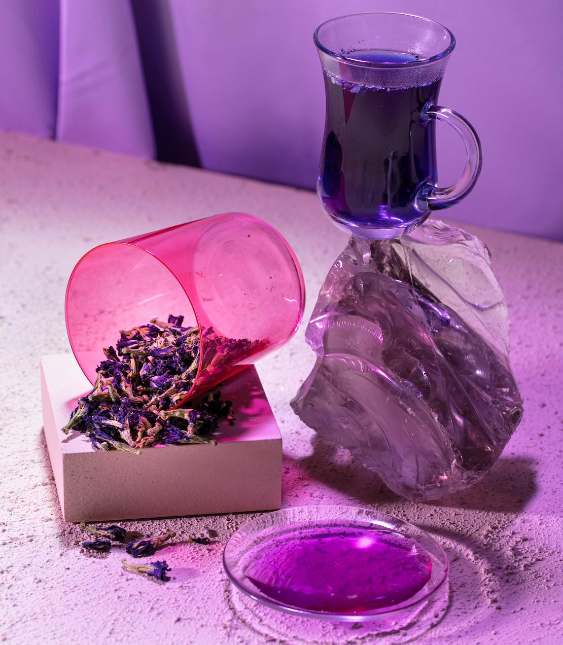 Butterfly pea flower tea in a glass, with dried flowers spilled from a pink cup on a textured surface.