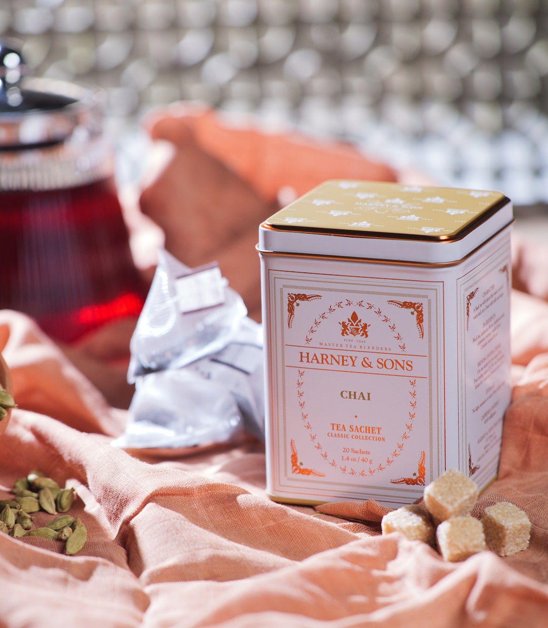 Classic tin of Harney & Sons chai sachets with a teapot, glass of chai, and spices on a textured fabric background.