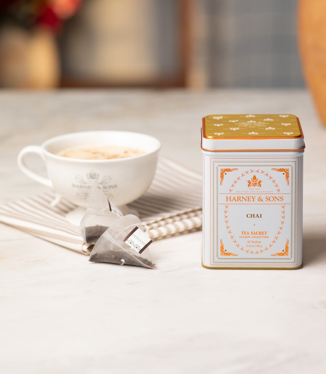 A tin of Harney & Sons Chai tea sits on a light marble surface next to a white teacup and two tea sachets.