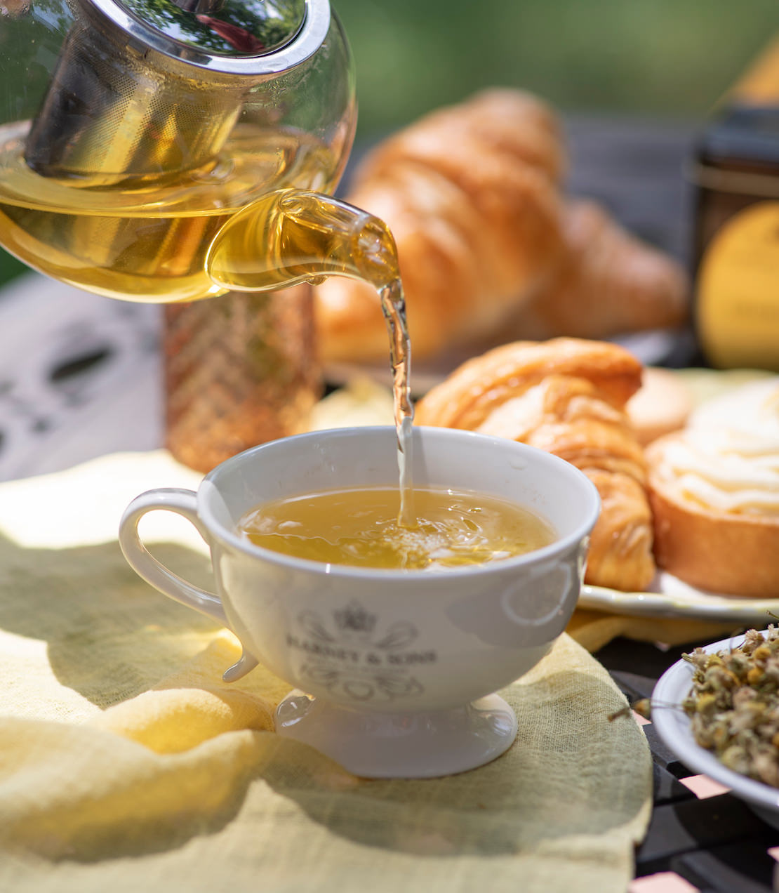 Chamomile tea being poured into a white teacup with pastries in the background.