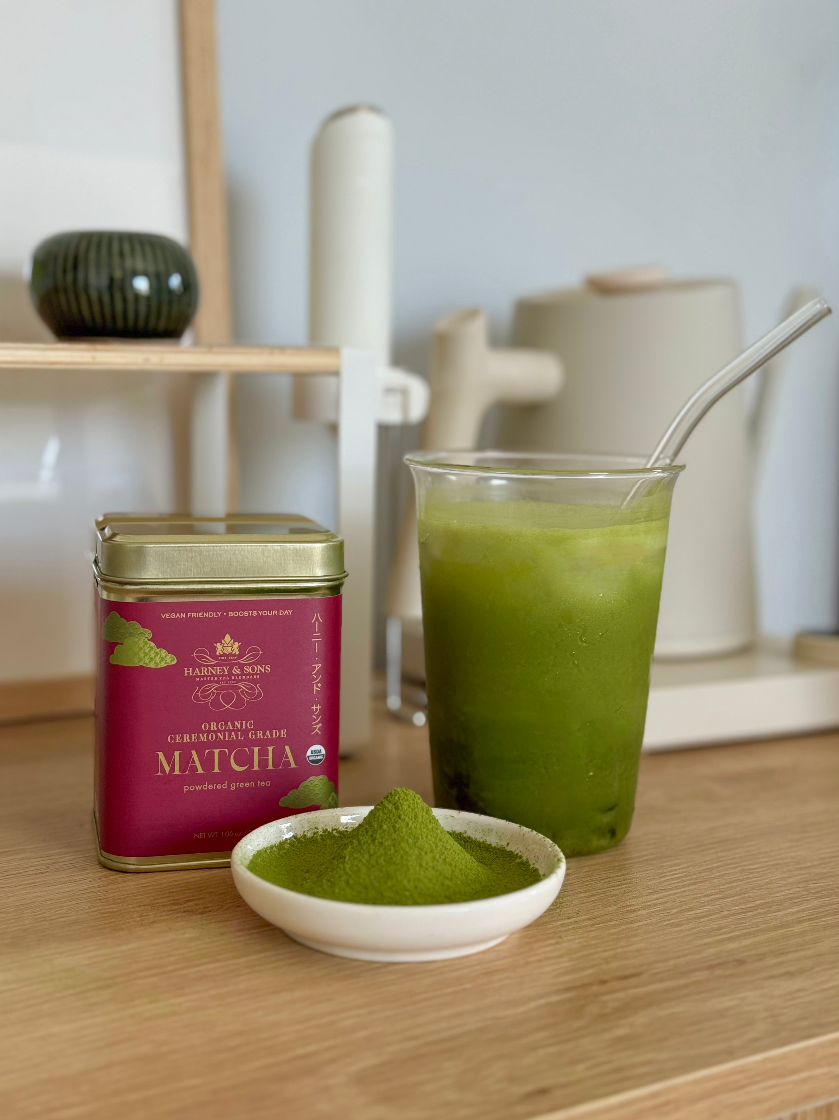 A tin of Matcha Green Tea Beans by The Matcha Society sits next to a glass of prepared green matcha beverage with a metal straw and a small white dish of loose matcha powder on a wooden surface.