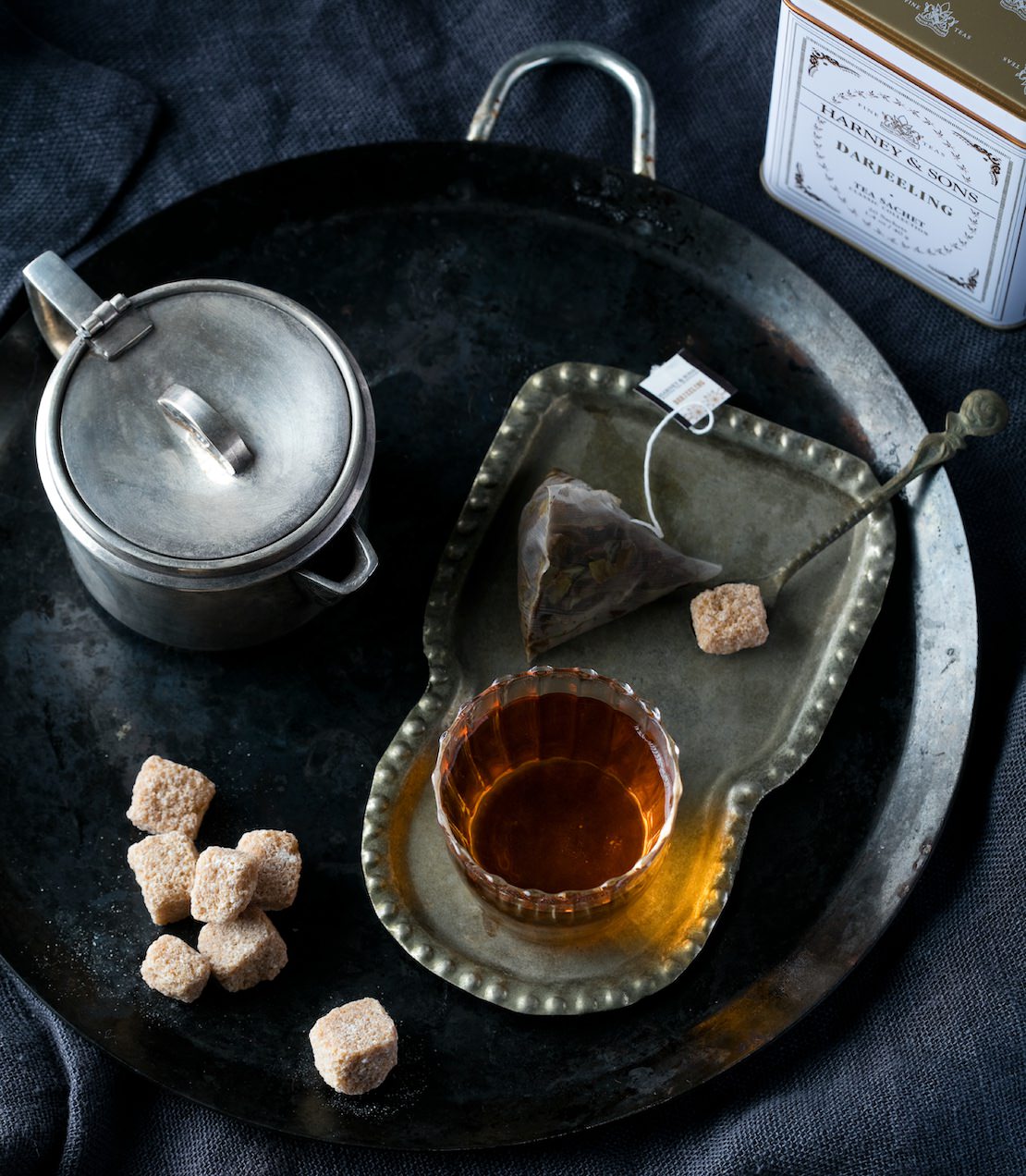 Tea set with cup, sugar cubes, and tea sachet on a dark surface next to a Darjeeling classic tin of 20 sachets.