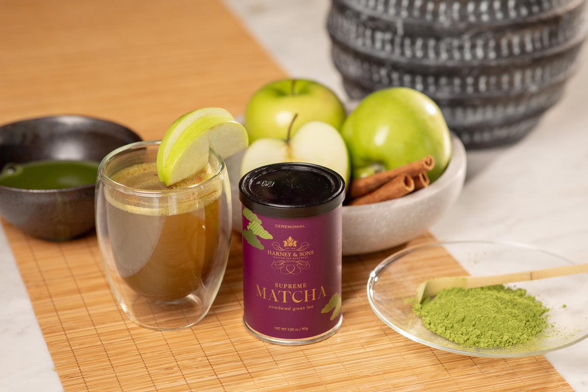 A can of Supreme Matcha tea is displayed on a bamboo mat next to a glass of prepared matcha tea with a lime wedge, a bowl of green apples, cinnamon sticks, and a small dish of loose matcha powder.
