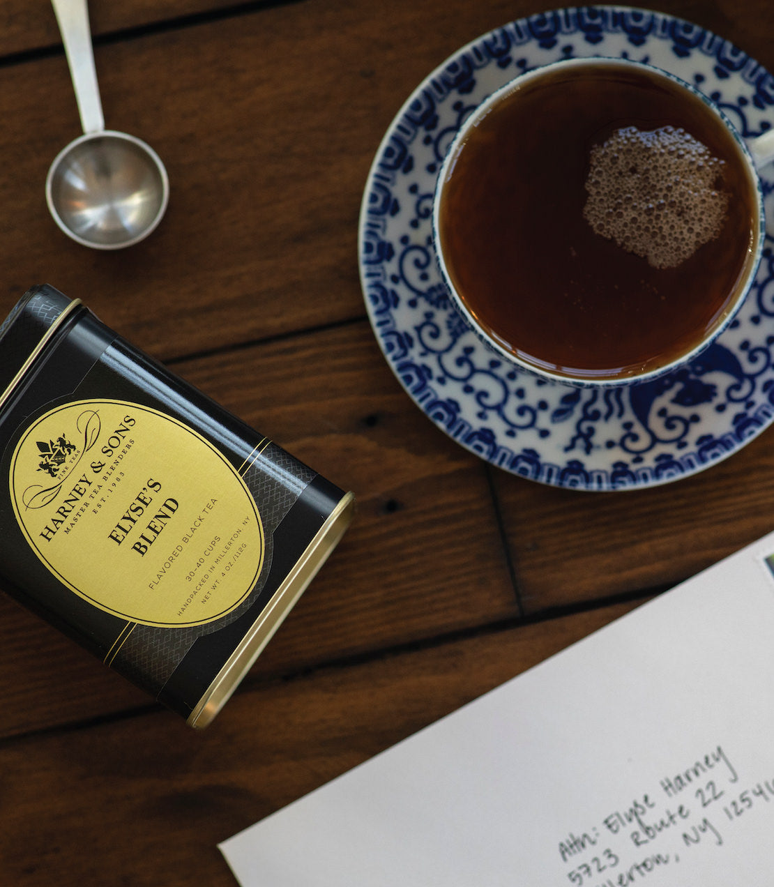 Elyse's Blend tea tin beside a cup of brewed tea on a wooden table, with a decorative blue and white plate.