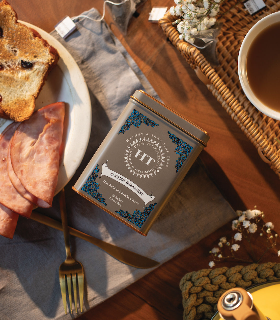 Tin of 20 English Breakfast tea sachets on a rustic table with toast and ham, showcasing a cozy breakfast setting.