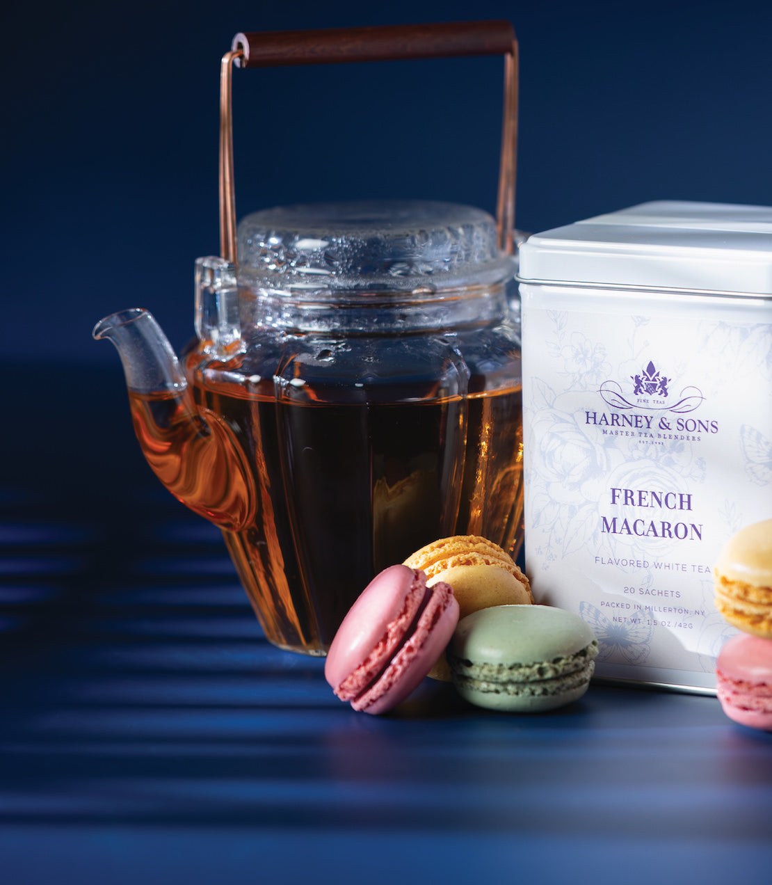Tin of 20 French Macaron tea sachets beside a glass teapot and colorful macarons on a blue background.