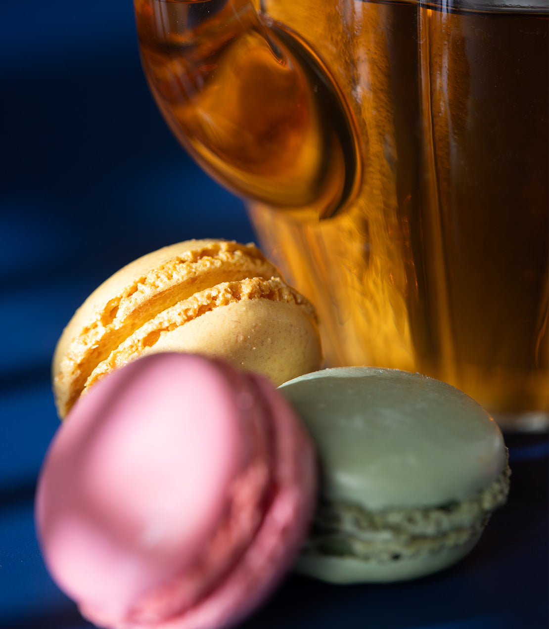 Colorful French macarons in pink, green, and yellow, with a glass of amber beverage in the background.