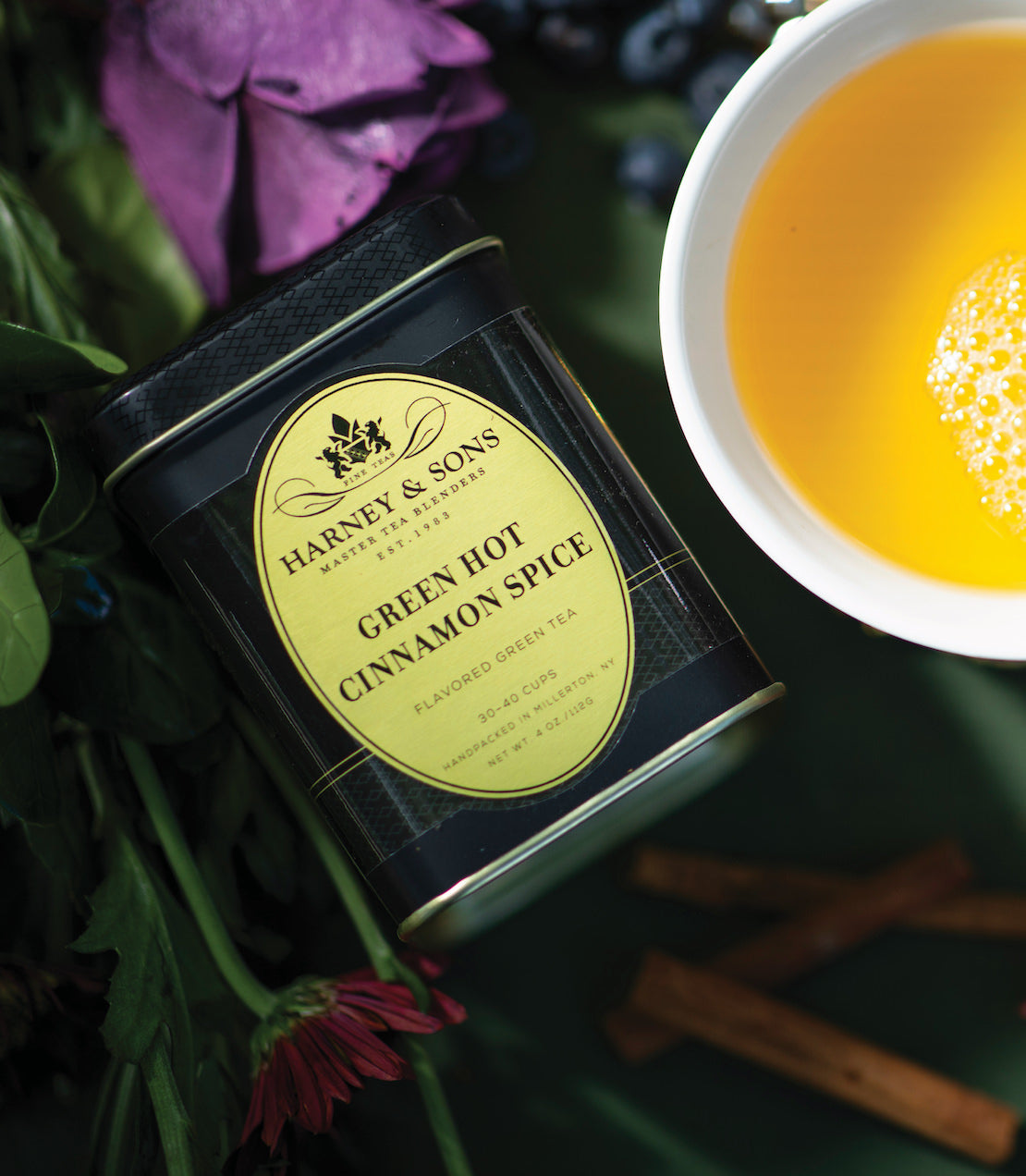 Harney & Sons Green Hot Cinnamon Spice tea canister beside a cup of brewed tea and cinnamon sticks.