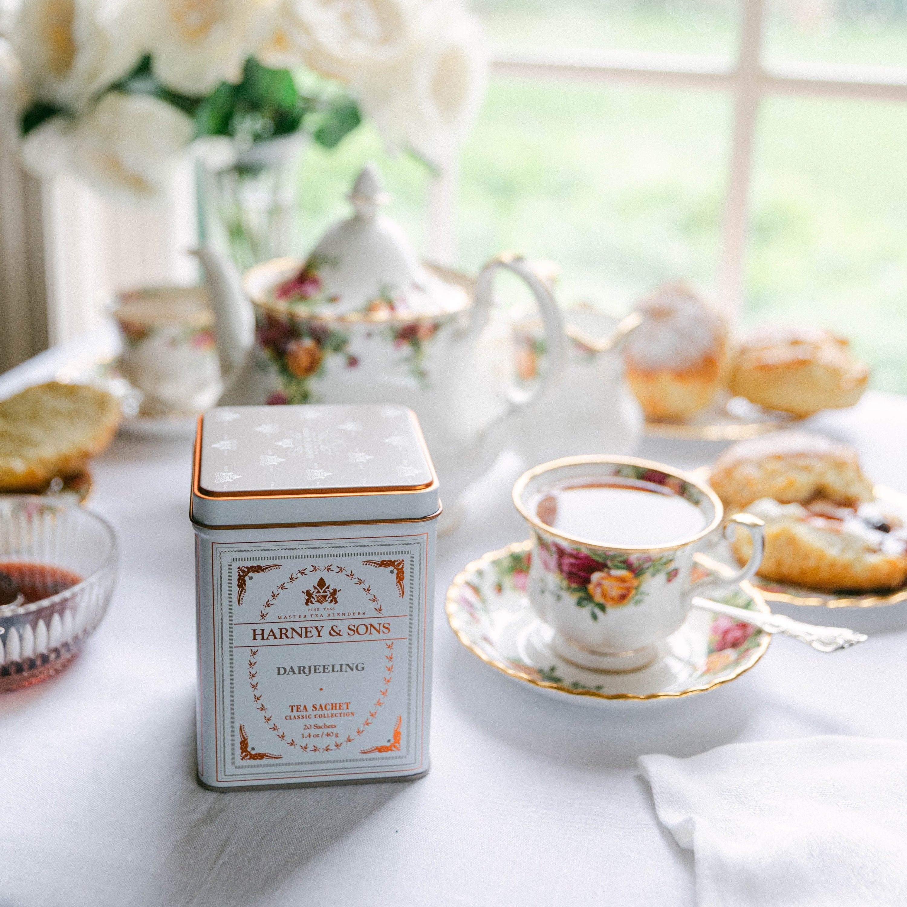A square white tea tin with gold detailing and floral illustrations sits on a white tablecloth next to a matching teacup and saucer in a bright, sunlit room with a window and flowers in the background.