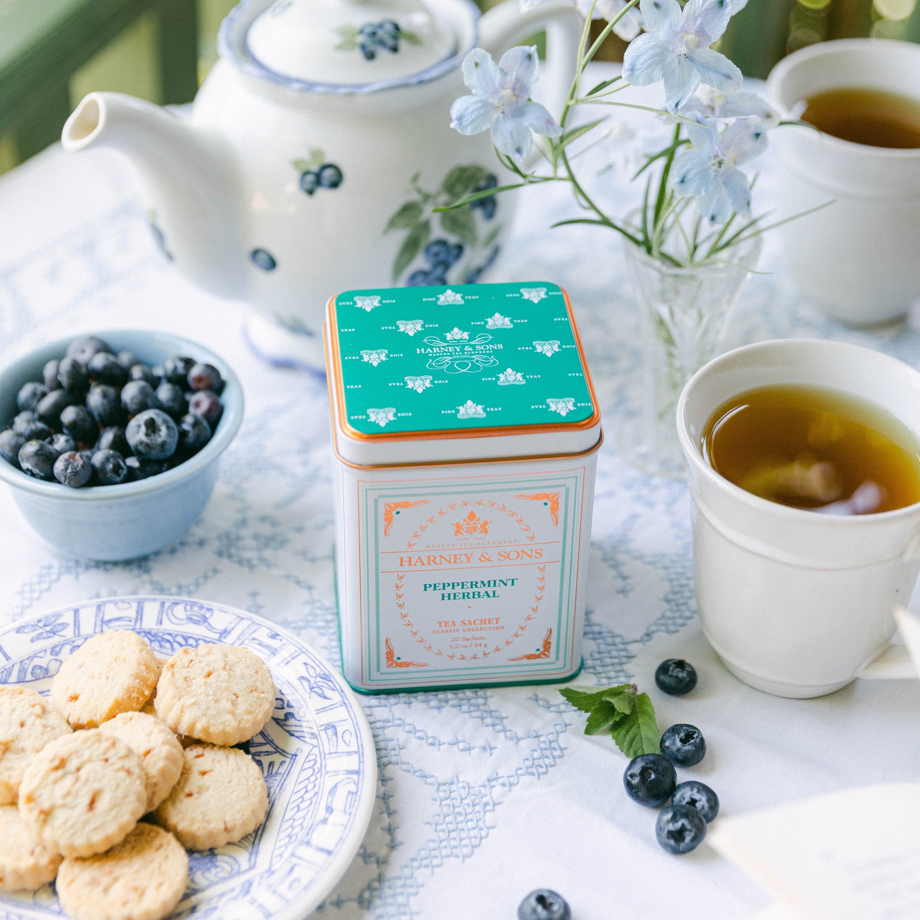 A square tin of Peruvian Blueberries sits on a table set for tea, surrounded by blueberries, cookies, a teapot, and cups on a blue-and-white tablecloth.