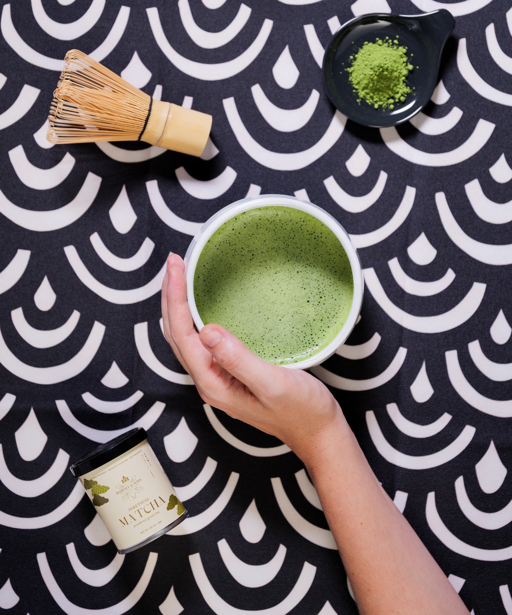 A hand holds a white cup filled with green matcha tea over a black and white patterned surface. A bamboo whisk, a small dish of green matcha powder, and a candle labeled 'MATCHA' are arranged around it in a styled flat-lay composition.