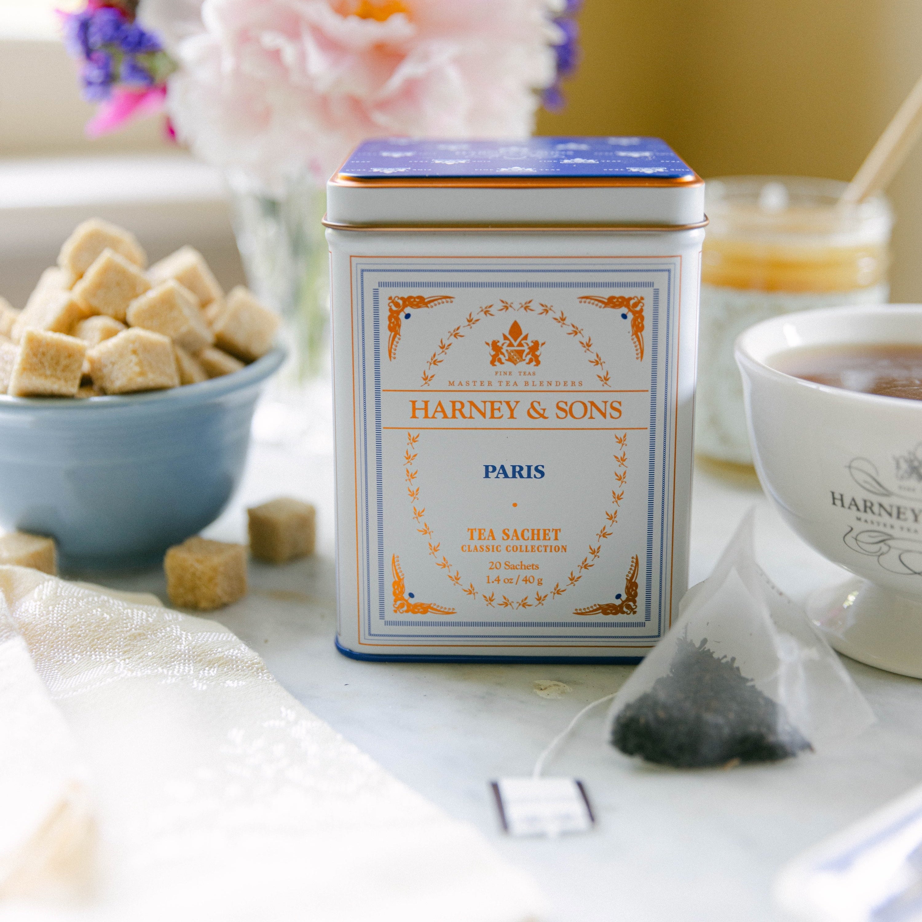 A tin of Harnet & Sons Paris Tea Secret tea sits on a tabletop with a teabag, a cup, sugar cubes, and flowers in the background.