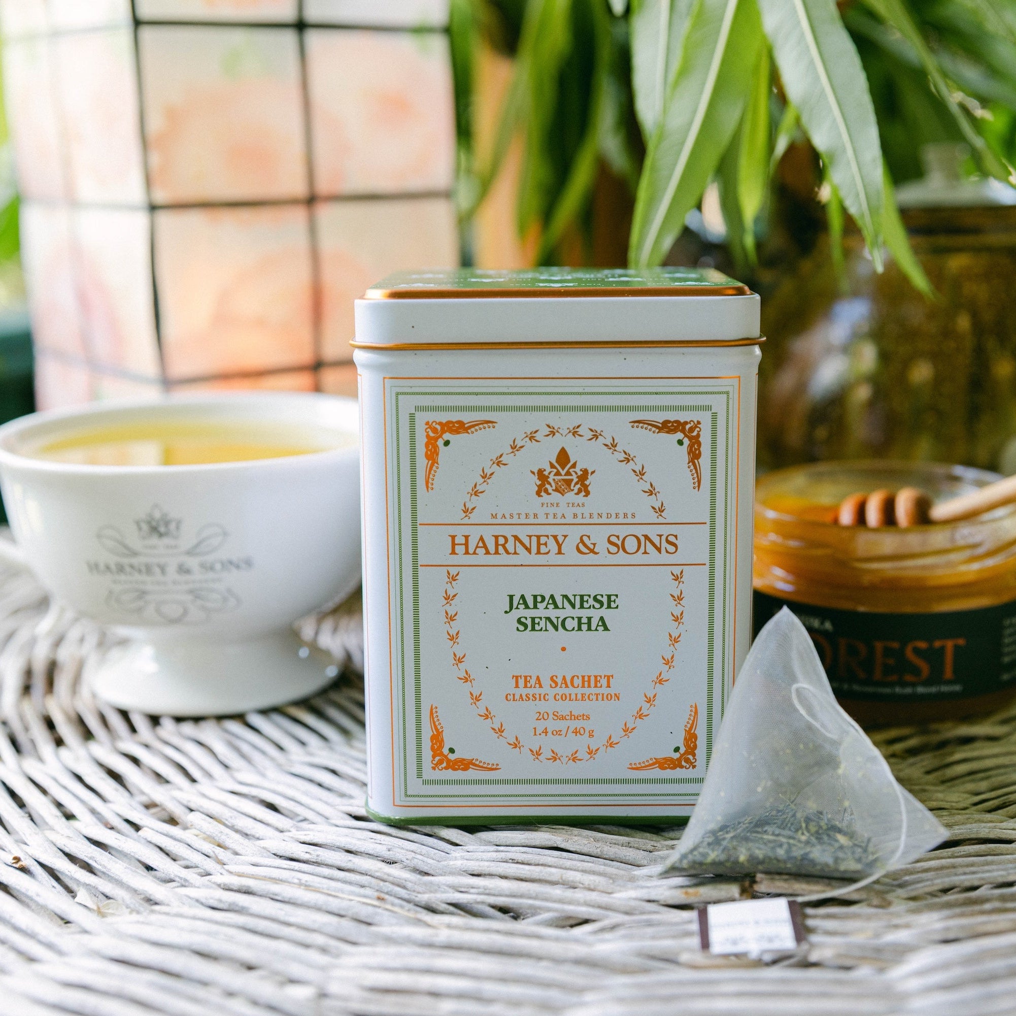 A tin of Harney & Sons Japanese Sencha tea stands on a woven table surface, with a teacup, honey pot, and cookies in a real outdoor setting in the background.