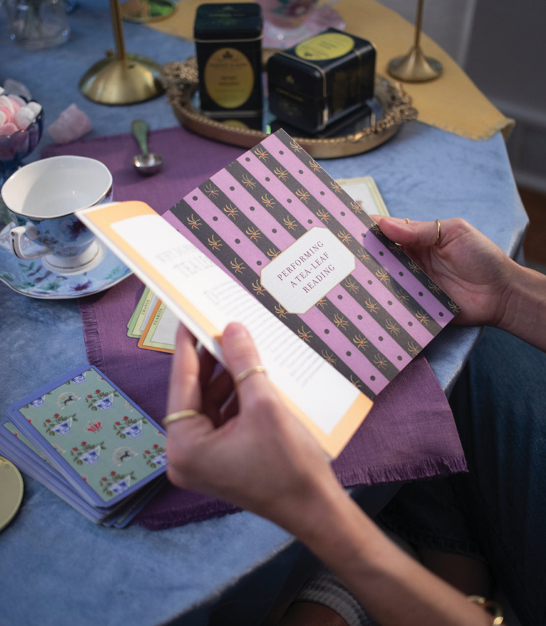 A person holds an open book titled "How to Read Tea Leaves" on a table set with tea and cards.