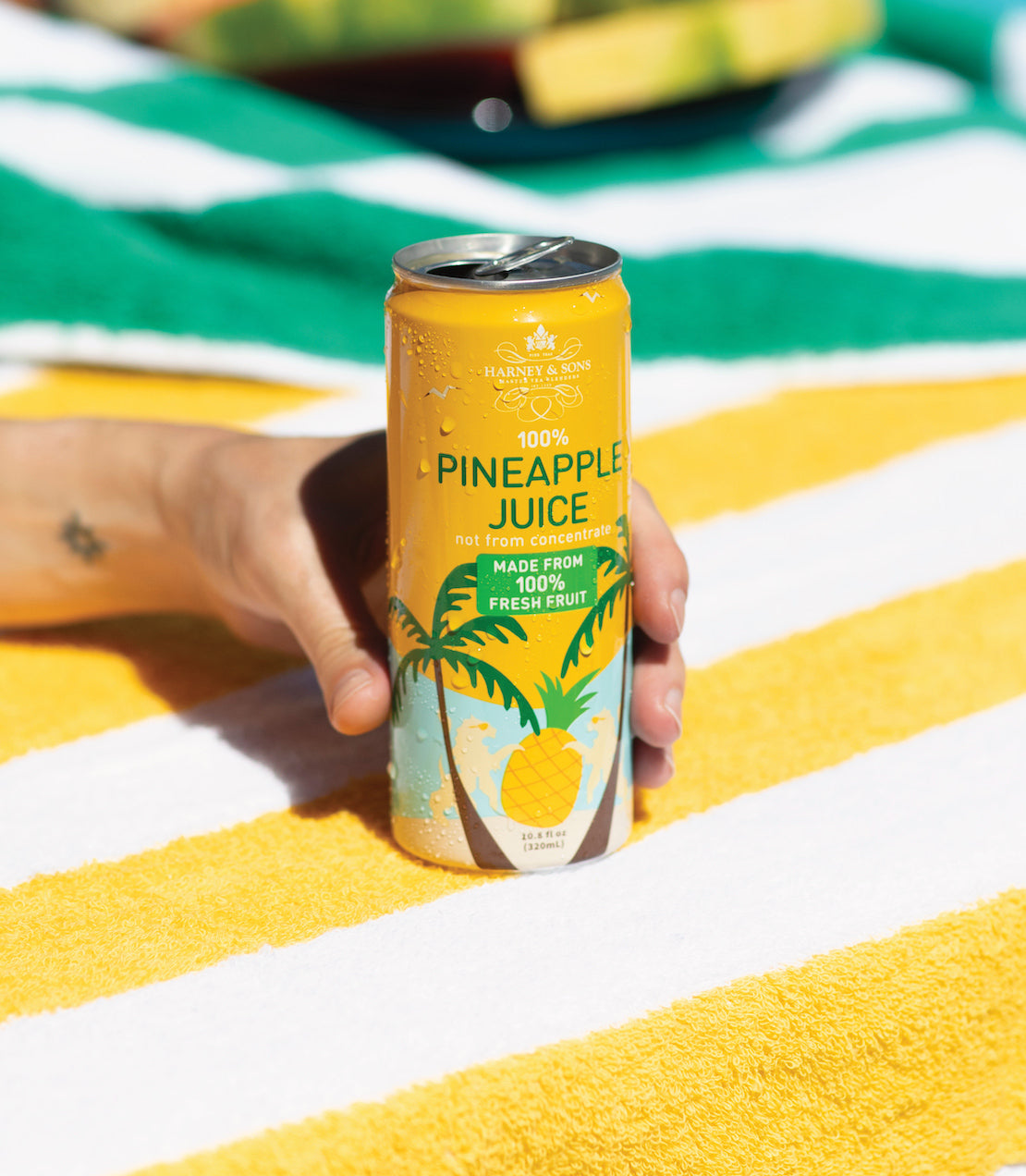 Can of Harney & Sons 100% Pineapple Juice held in hand, featuring vibrant tropical design on a striped beach towel.