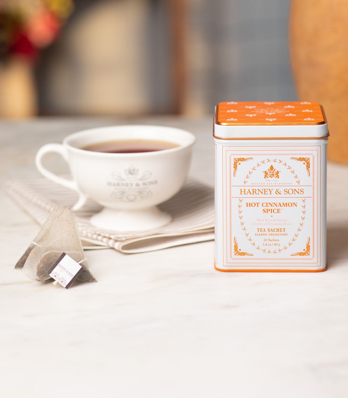 A tin of Harney & Sons Hot Cinnamon Spice tea sits on a light marble surface next to a white teacup with tea and two tea sachets.