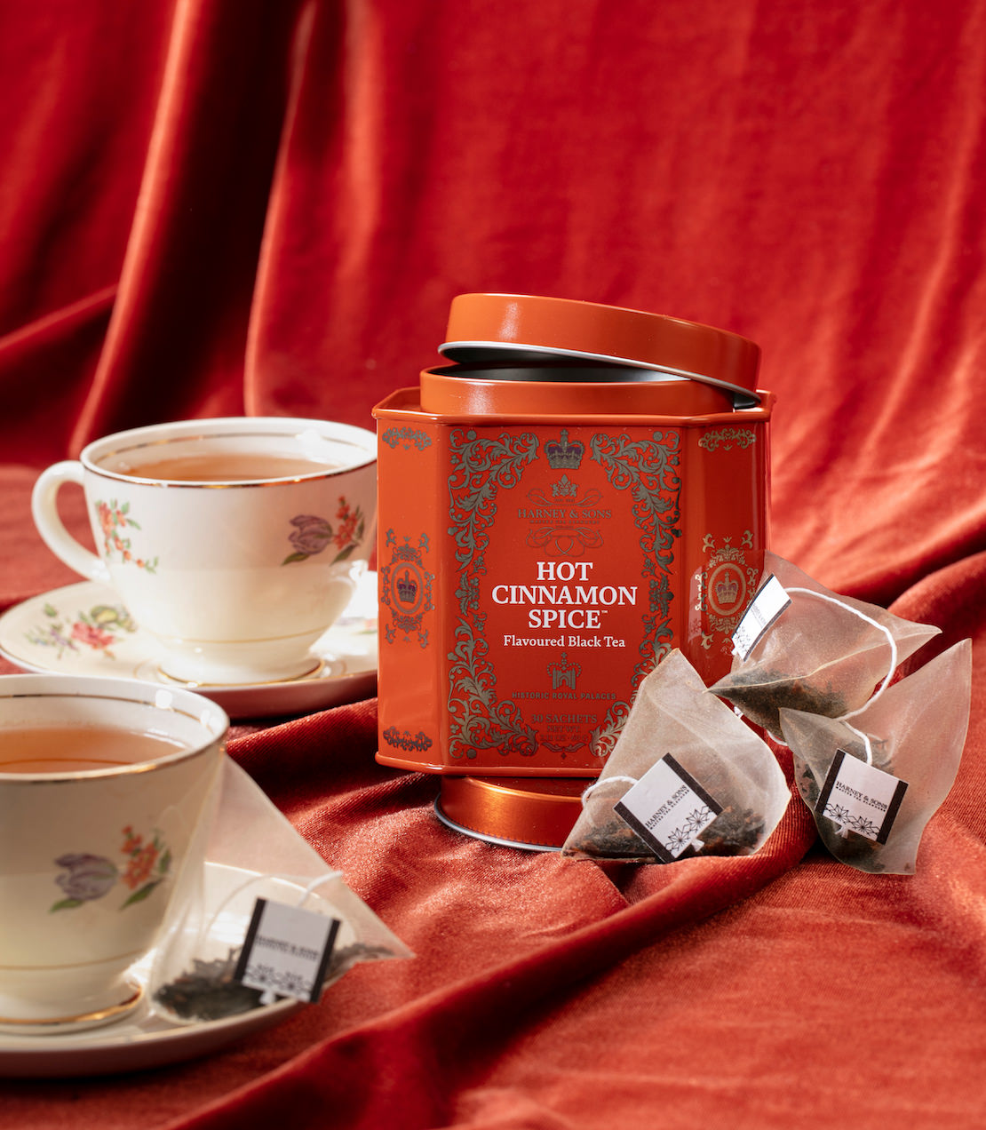Hot cinnamon spice tea with teacups, tea canister, and tea sachets on a red fabric background.