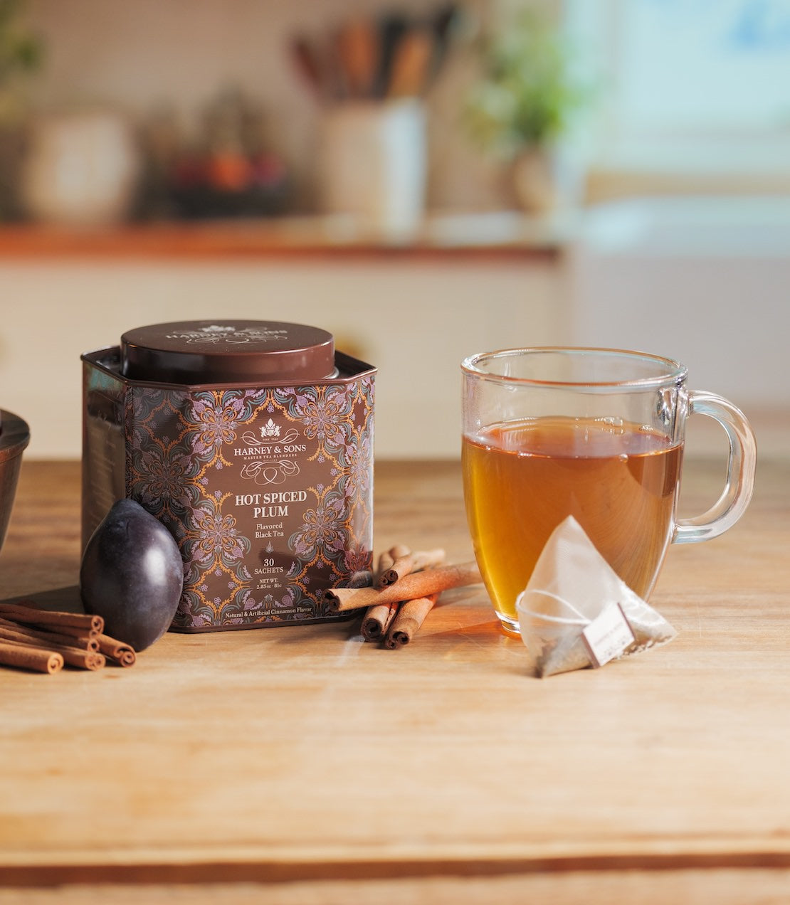 A tin of Hot Spiced Plum tea sits on a wooden table next to a glass mug filled with amber tea, a tea bag, cinnamon sticks, and a plum in a softly blurred kitchen setting.