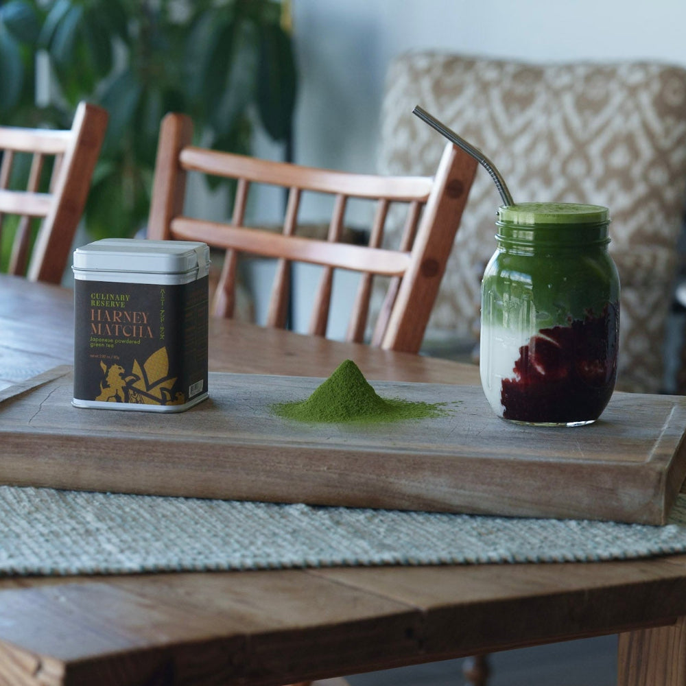 A wooden tray on a dining table holds a small black tin labeled Harney & Sons, a pile of green powder, and a glass jar filled with layered green and red drink and topped with a metal spoon. The scene is set in a real indoor environment with chairs and plants in the background.