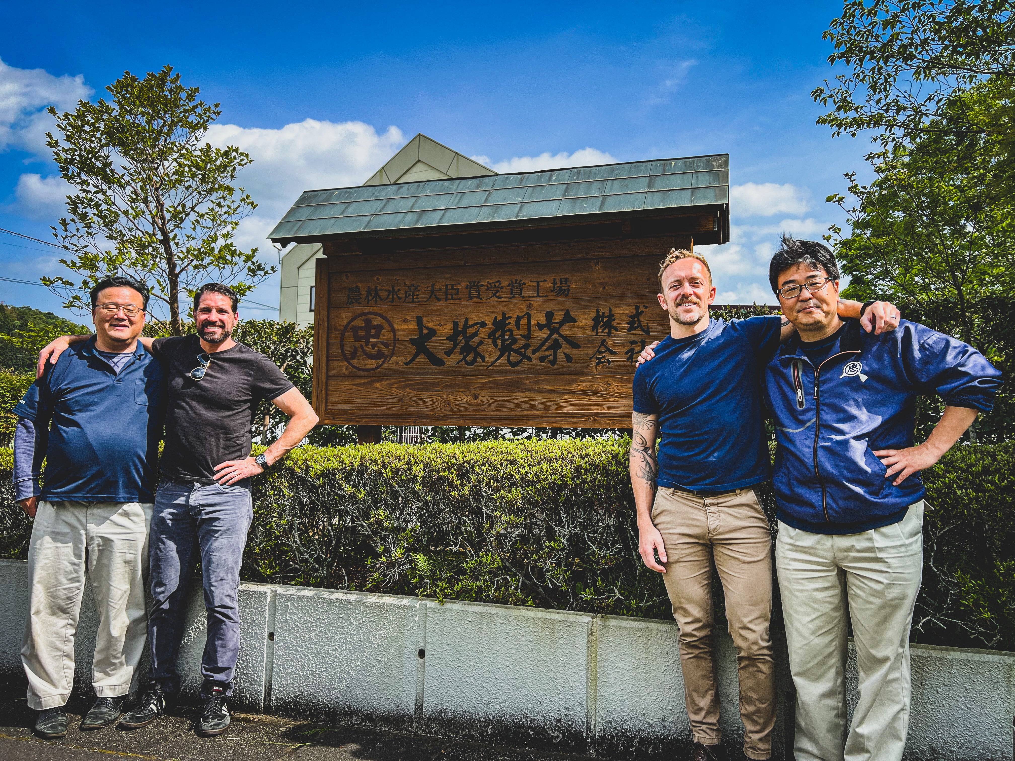Group of four men posing in front of a wooden sign for Ichiban Sencha tea factory, surrounded by greenery.