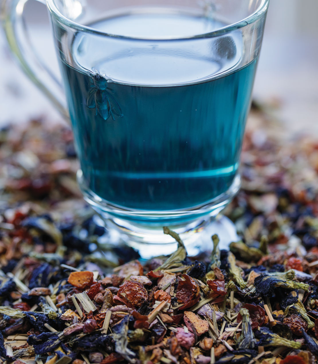 A glass of Indigo Punch tea surrounded by colorful dried herbs and flowers, showcasing its vibrant blue hue.