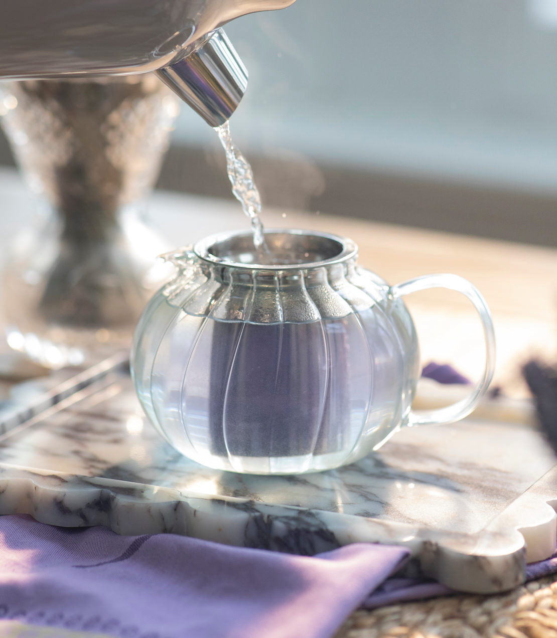 Glass teapot being filled with water on a marble surface.