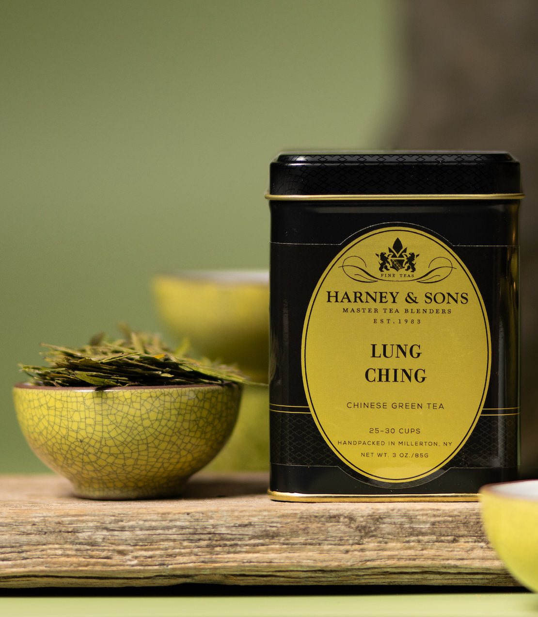 Harney & Sons Lung Ching Chinese Green Tea tin with tea leaves in a bowl on a wooden surface.