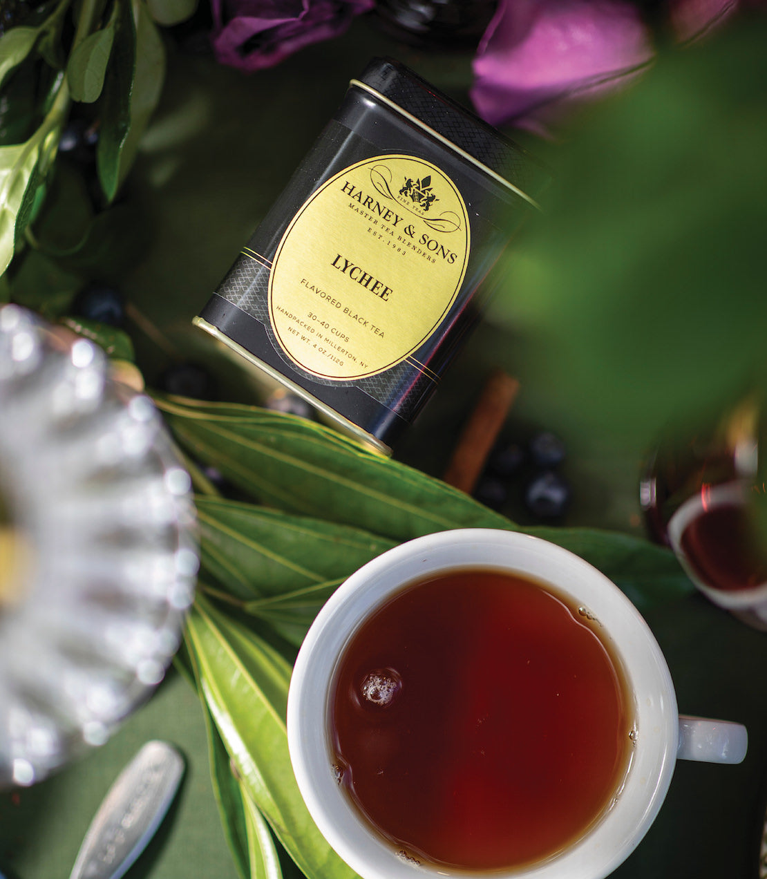 A cup of brewed lychee black tea beside a tin of Harney & Sons lychee tea, surrounded by green leaves and berries.