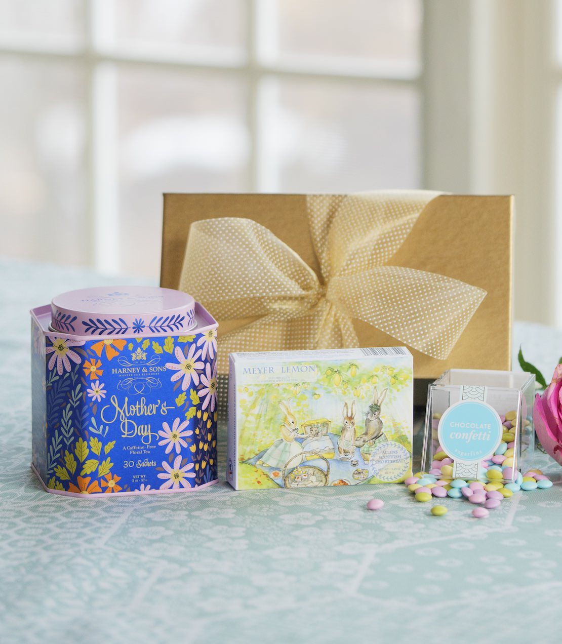 Mother's Day Tea Tin, Meyer Lemon Shortbread, and Chocolate Confetti candy cube on a table with a gold gift box with ribbon in the background.