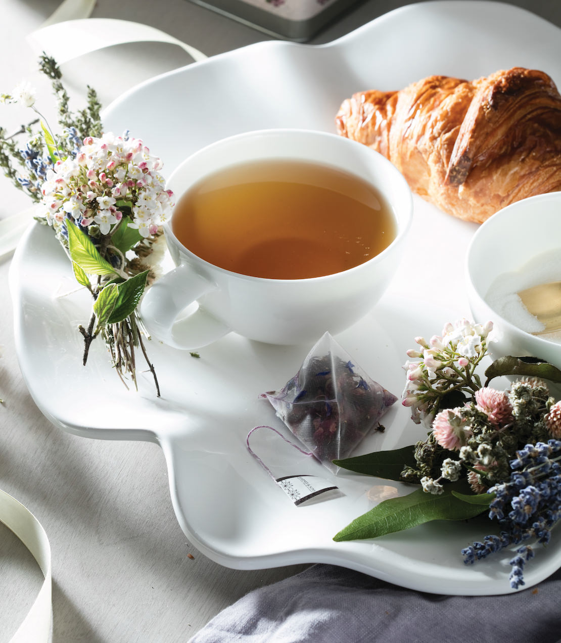 A white tray with a cup of tea, a croissant, and a sachet of tea surrounded by fresh flowers for Mother's Day.