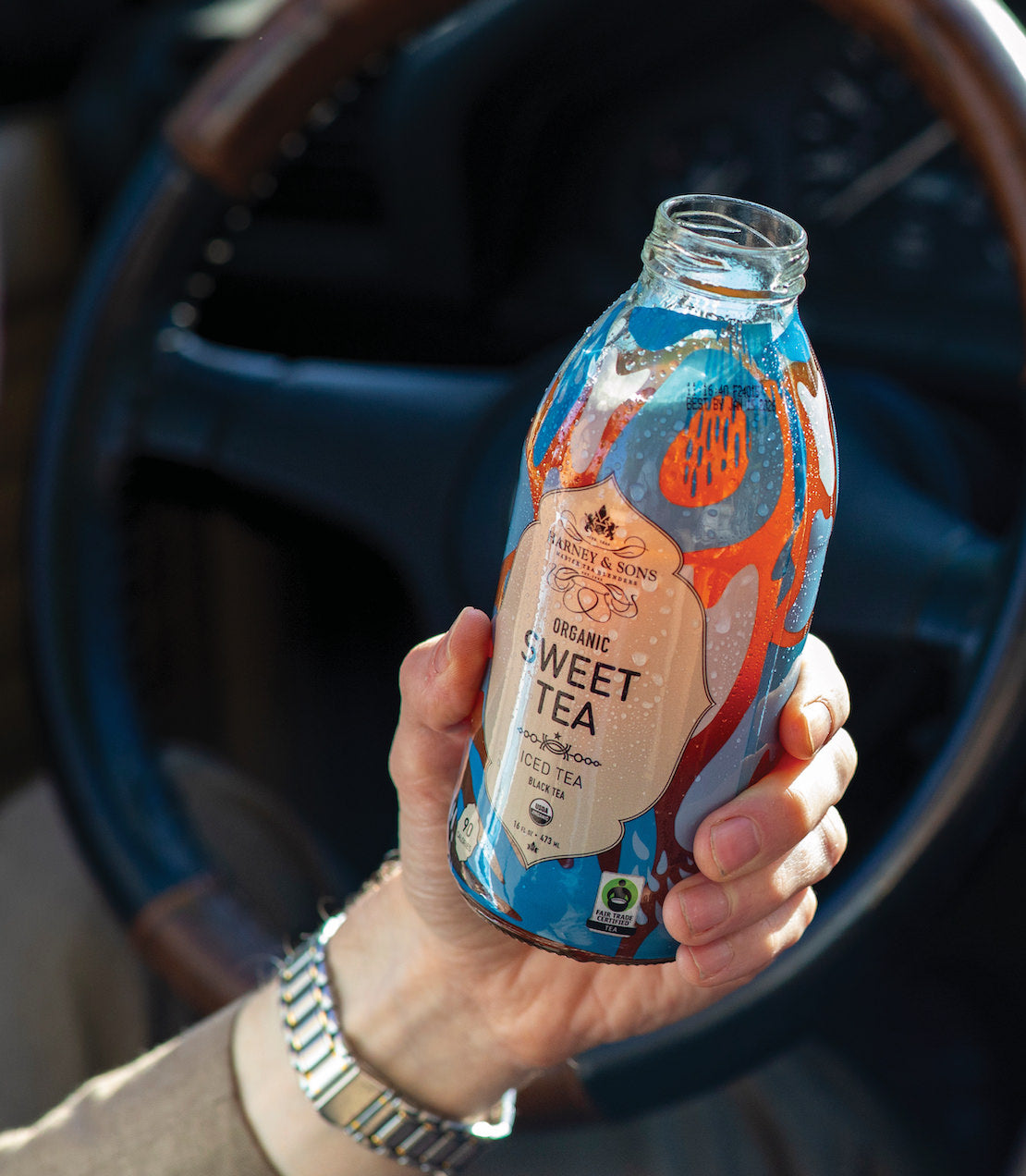 Hand holding a bottle of Organic Black Sweet Iced Tea with a colorful label, set against a car interior background.