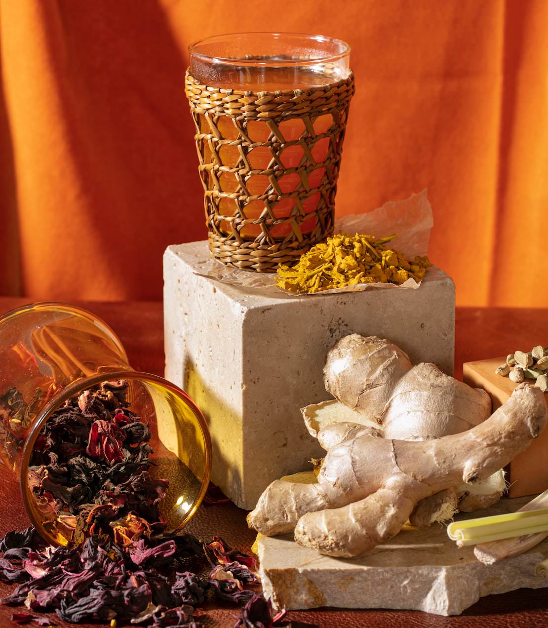 A glass of organic ginger turmeric tea surrounded by ginger root, dried herbs, and vibrant orange backdrop.